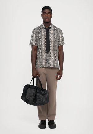 Homme portant une chemise à imprimé serpent à manches courtes avec une cravate noire, un pantalon beige, des chaussures noires, tenant un sac de voyage noir à motifs.