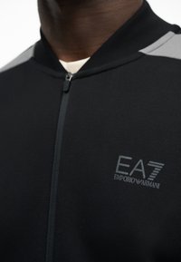 Κοντινό πλάνο ενός ατόμου που φορά ένα μαύρο μπουφάν με φερμουάρ, με γκρίζες λεπτομέρειες στους ώμους και λογότυπο EA7 Emporio Armani στο στήθος.