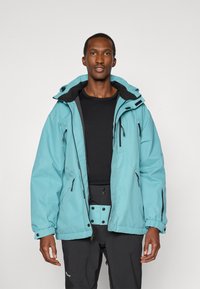 Türkise wasserdichte Jacke mit schwarzen Akzenten, Kapuze und mehreren Reißverschlusstaschen; getragen über einem schwarzen Shirt und kombiniert mit schwarzen Hosen.