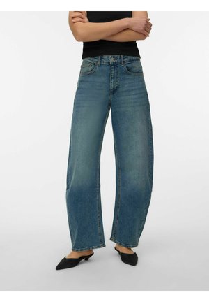 Persona che indossa jeans blu a gamba larga a vita alta con cinque tasche e scarpe nere a punta con tacco basso, con le braccia incrociate sopra una maglietta nera.