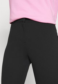 Närbild av en person som bär svarta åtsittande byxor med framdragkedja och en instoppad rosa topp.