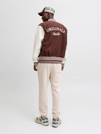 Bruine en crèmekleurige varsityjas met de tekst "ORIGINALS Studio", gecombineerd met crèmekleurige joggers en beige sneakers. Heeft geribbelde manchetten en onderkant.