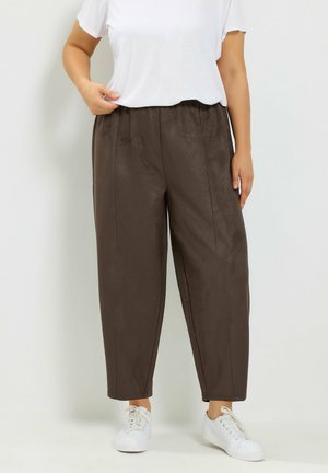 Donna che indossa pantaloni marroni ampi, top bianco a maniche corte e sneakers bianche, in piedi davanti a uno sfondo chiaro e semplice.