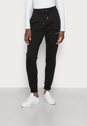 Personne portant un pantalon de jogging noir avec une taille à cordon et des baskets blanches, debout devant un fond clair uni.