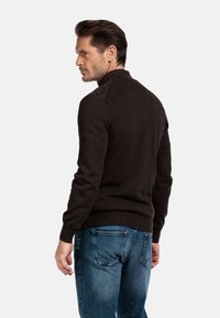 Donkerbruine trui van zacht materiaal met een hoge kraag. Voorzien van schouderaccenten en ribbelmanchetten, gecombineerd met blauwe jeans.