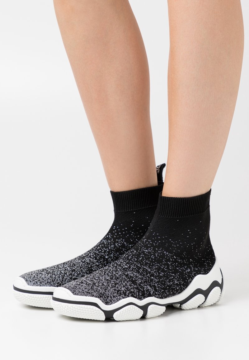Baskets chaussettes noires et blanches avec dessus en maille texturée et semelle blanche épaisse portées sur jambes nues sur fond blanc.