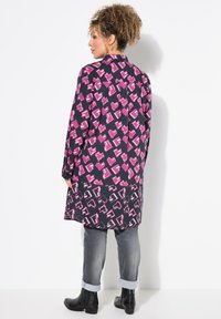 Langes schwarzes Hemd mit pinken abstrakten Mustern; verfügt über einen Kragen, Knopfmanschetten und eine lässige Passform, kombiniert mit grauen Jeans und schwarzen Stiefeletten.