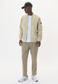Chaqueta bomber beige con cremallera frontal, pantalones beige a juego, camiseta blanca y zapatillas blancas. La gorra tiene un pequeño logo.