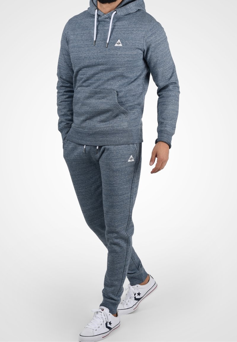 Mann in blauer melierter Kapuzenjacke und passenden Jogginghosen mit weißen Sneakern, steht mit einer Hand in der Tasche und einer entspannt.