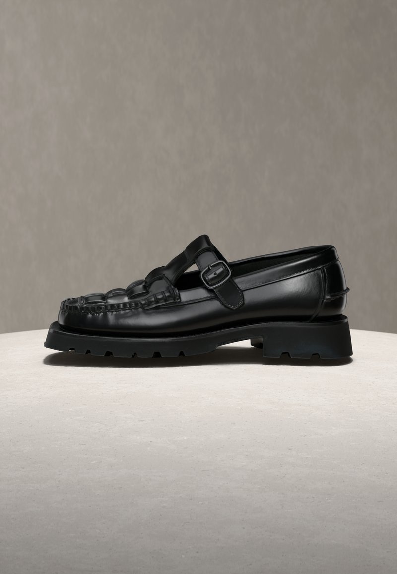 Mocassin en cuir noir avec semelle en caoutchouc épaisse et bride à boucle réglable sur fond et surface neutres.