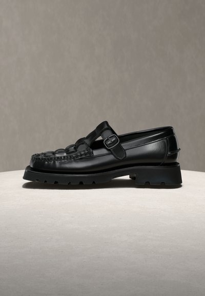 Mocassin en cuir noir avec semelle en caoutchouc épaisse et bride à boucle réglable sur fond et surface neutres.