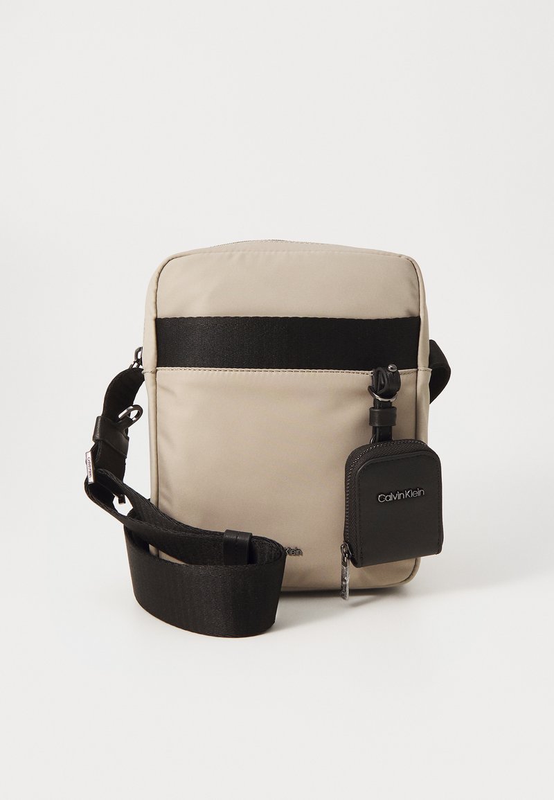 Borsa a tracolla beige con una tracolla nera, dotata di una banda orizzontale nera frontale e di una piccola tasca con zip nera attaccata. Tessuto dal tocco liscio.