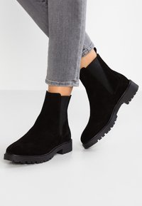 Persona con botas de ante negras y suelas de engranaje, usando jeans grises recortados, de pie sobre un suelo blanco.