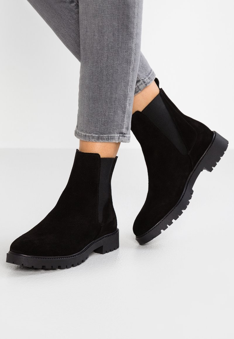 Persona con botas de ante negras y suelas de engranaje, usando jeans grises recortados, de pie sobre un suelo blanco.