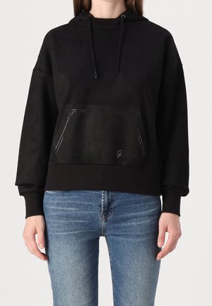 Femme portant un sweat à capuche noir avec une poche avant et des cordons, associée à un jean bleu, debout devant un fond clair uni.