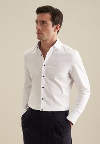 Witte button-up shirt met zwarte knopen, gestructureerde kraag en contrasterende binnenkraagdetails; gecombineerd met donkere broeken.