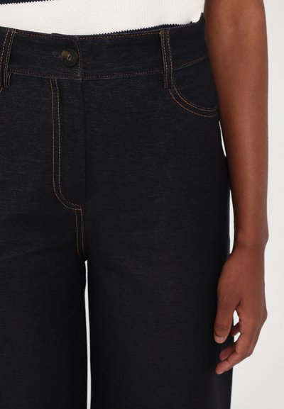 Tamsiai mėlynos džinsinės kelnės su oranžinėmis siūlėmis. Su mygtukų uždarymu, priekinėmis kišenėmis ir tiesiomis kojomis. Matoma minkšta tekstūra.