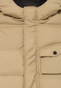 Beige vadderad jacka med hög krage, med knäppe och en bröstficka. Tyget har en slät, något strukturerad yta.