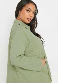 Olivgrön oversize-jacka med krage, två framfickor och knapppaneler på axlarna. Tillverkad av slitstark väv.