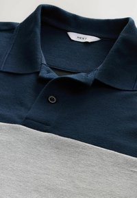 Tummansininen ja vaaleanharmaa raidoitettu polo-paita, jossa on ribbitekstuuria, kolme napin aukkoa ja kauluksessa näkyvä "NEXT"-merkillä varustettu etiketti.