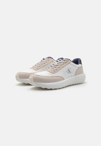 Beige en witte sneakers met een gladde textuur en een dikke witte zool. Voorzien van een logodetail en contrasterende marineblauwe accenten op de hiel en de tong.