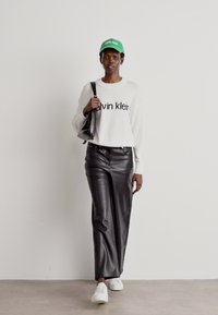 Balts džemperis ar "Calvin Klein" logo, melnas ādas platās bikses, zaļš cepure, un baltas sporta apavi, papildināti ar melnu roku somu.