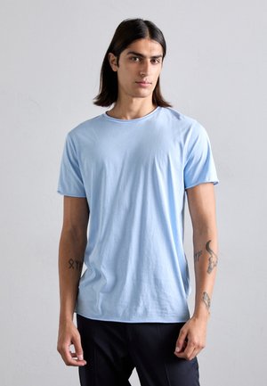Mann mit langen dunklen Haaren trägt ein hellblaues T-Shirt mit kurzen Ärmeln und schwarze Hosen, steht vor einem schlichten hellgrauen Hintergrund.