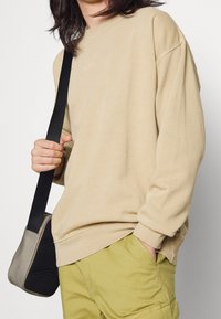 Sudadera beige de cuello redondo con corte relajado, puños acanalados y aberturas laterales, combinada con pantalones cortos verde oliva y un pequeño bolso mensajero negro.