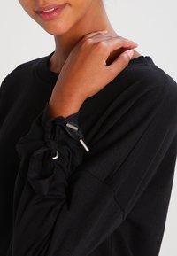 Prenda negra de manga larga con detalles fruncidos en los puños, que cuenta con lazos ajustables y acentos de metal. Textura de tela suave.