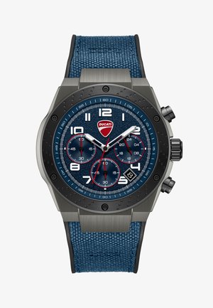 Kronografska ura z modrim teksturiranim številčnico, belimi številkami, rdečimi poudarki, tremi podštevilčnicami, datumskim okencem ob 4. uri in modrim platnenim paščkom.