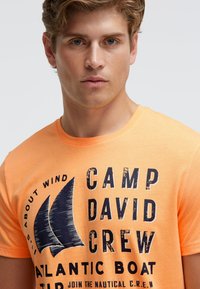 Orange cotton t-shirt featuring a graphic print of sailboat designs and text in dark blue. Crew neck and short sleeves.