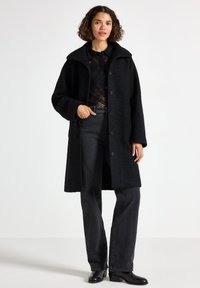 Svart teddyjacka med hög krage och framknäppning, parat med mörka jeans och svarta stövlar. Strukturerad tyg med en lös passform.