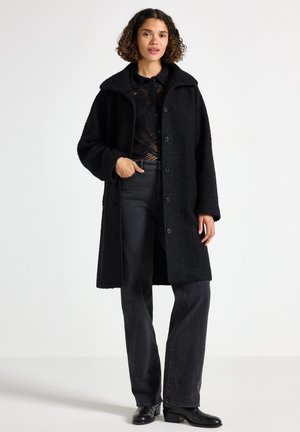 Svart teddyjacka med hög krage och framknäppning, parat med mörka jeans och svarta stövlar. Strukturerad tyg med en lös passform.