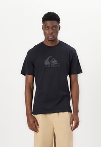Black cotton t-shirt with a crew neck and short sleeves, featuring a textured gray logo in the center. Worn with beige shorts.