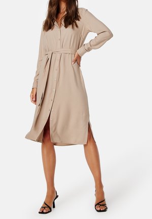 Vrouw draagt een beige jurk met lange mouwen, knopen, ceintuur, zijslits en zwarte sandalen met hoge hakken en bandjes, poserend met hand op de heup.