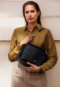 Schwarze Lederhandtasche mit glatter Textur und goldenen Hardware. Verfügt über einen Schultergurt und ein dezentes goldenes Logo. Das Modell trägt ein grünes Shirt.