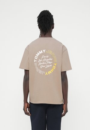 Férfi dreadlockkal, bézs színű pólót visel, amelyen a "Tommy Jeans" és városnevek körkörös mintában vannak nyomtatva a háton, fekete nadrág.