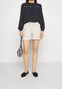 Blouse bleu marine à manches longues avec fermeture à boutons et surpiqûres décoratives, associée à un short beige clair à cordon de serrage et des chaussures plates noires.