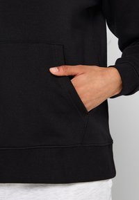 Czarna bluza dresowa z gładką fakturą, z kangurzą kieszenią i otwartym szwem na dole. Ręka umieszczona w kieszeni.