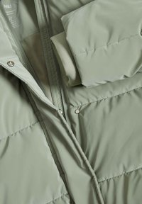 Lys grøn dunjakke med en glat, mat overflade. Den har en lynlåslukning og trykknapper. Indersiden viser en foldet ærme i matchende farve.