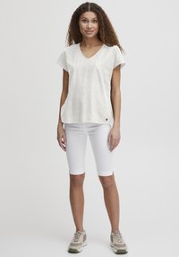 Blusa in lino bianco leggera e a maniche corte con scollo a V e spacchi laterali, abbinata a pantaloni bermuda bianchi aderenti e sneaker beige.