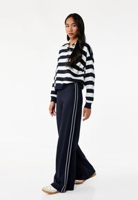 Navy og hvid stribet langærmet polo-shirt parret med navy vide bukser med hvide sidestriber, suppleret af hvide sneakers.