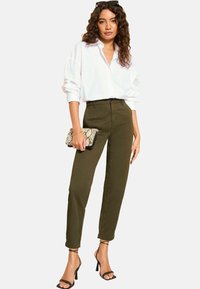 Pantalons taille haute vert olive, coupe ajustée, assortis à une chemise blanche ample. Accessoires avec une pochette à motif serpent et des talons noirs.