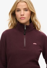 Bordowy polarowy golf z wysokim kołnierzem, ćwierć zamkiem i małym logo na klatce piersiowej. Charakteryzuje się miękką fakturą i luźnym krojem.