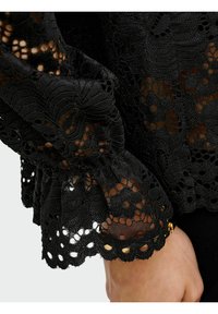 Manica in pizzo nero con bordi scallopati, intricate fantasie floreali e polsino arricciato su un polso che indossa un bracciale dorato.