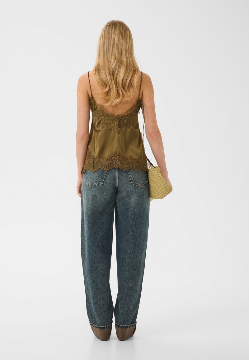 Femme aux longs cheveux blonds portant un caraco vert olive bordé de dentelle, un jean bleu ample, des chaussures marron, tenant une pochette tissée beige.