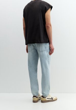 Mann trägt ärmelloses schwarzes Shirt, hellblaue Jeans und weiße Sneakers und steht vor einer schlichten hellgrauen Wand.
