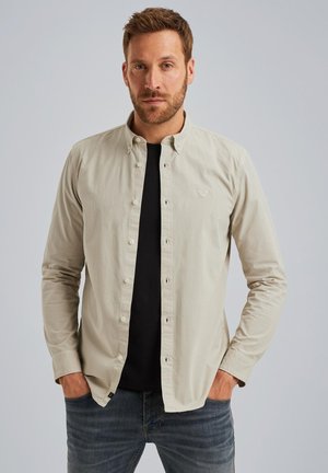 Chemise boutonnée beige clair avec un col structuré, des manches longues et un logo discret sur la poitrine, portée par-dessus un t-shirt noir.