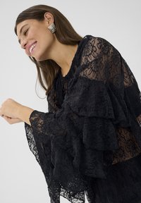 Melns mežģīņu tops ar kārtainām volānām, caurspīdīgu faktūru un apaļu izgriezumu. Modelim sudraba ģeometriskas auskari un viļņaini mati.