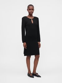 Femme portant une robe noire longueur genou avec des manches longues et un décolleté en forme de trou de serrure, associée à des ballerines Mary Jane noires pointues.